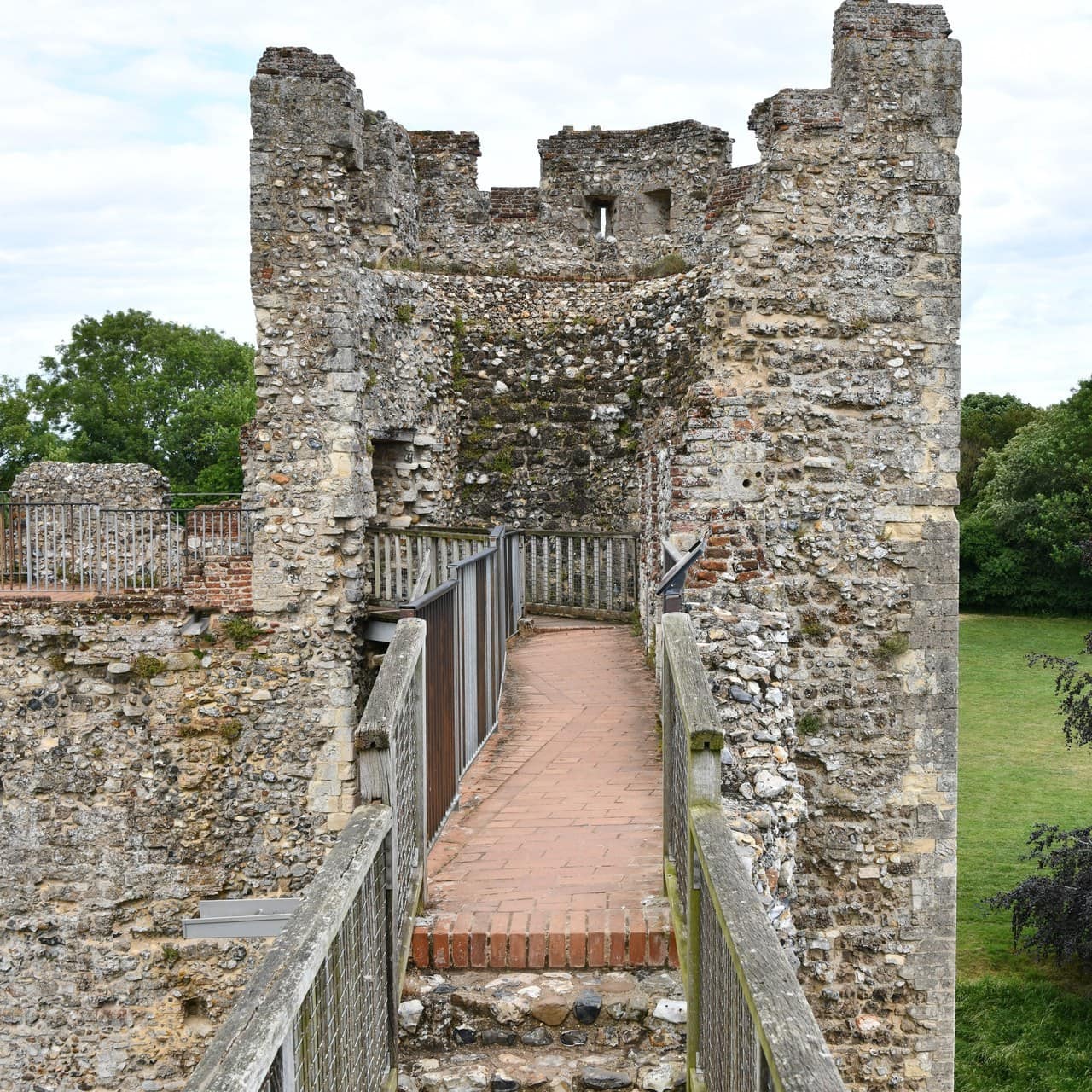 A square castle tower seen from the wall-walk. Its side walls and crenellated head stand, and the back of the tower is open so that the view carries straight through it. A walkway paved in red brick, carried on a modern deck with metal railings, runs from the foreground directly through the opening in the tower. The masonry is flint and rubble with dressed stone at the angles. Green countryside shows on either side. A square castle tower seen from the wall-walk. Its side walls and crenellated head stand, and the back of the tower is open so that the view carries straight through it. A walkway paved in red brick, carried on a modern deck with metal railings, runs from the foreground directly through the opening in the tower. The masonry is flint and rubble with dressed stone at the angles. Green countryside shows on either side.