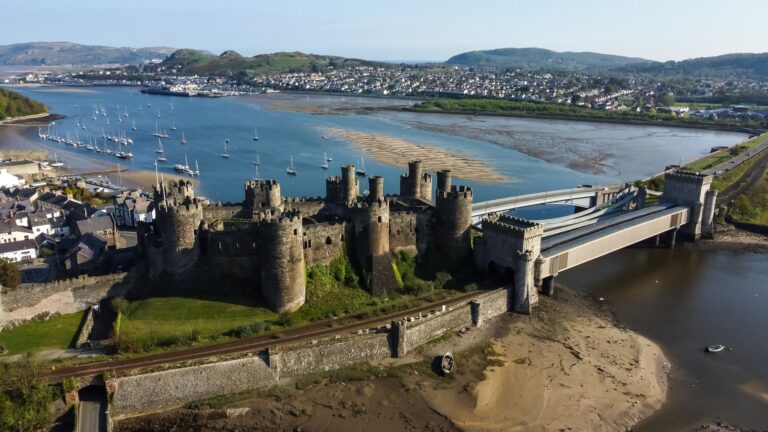 Aerial view of Conwy Castle on its rock beside the Conwy estuary, with the road and railway bridges running from its end across the river, moorings in the estuary and houses on the far shore beyond