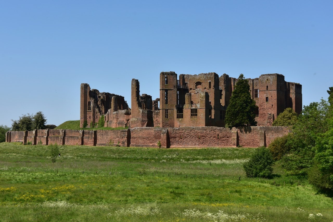 The red sandstone ruins of Kenilworth Castle rising behind a long, low outer curtain wall, seen across a meadow of long grass and wildflowers under a clear blue sky. The red sandstone ruins of Kenilworth Castle rising behind a long, low outer curtain wall, seen across a meadow of long grass and wildflowers under a clear blue sky.