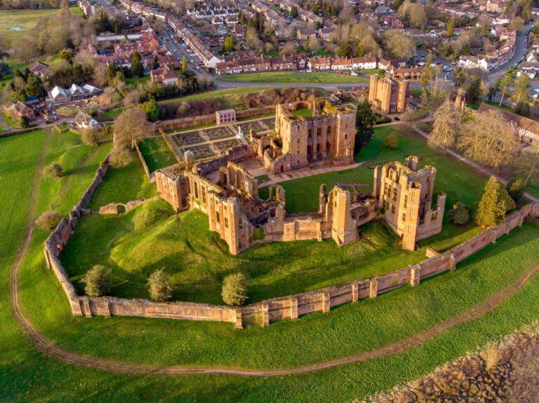 Home 5 Aerial view of the red sandstone ruins of Kenilworth Castle in low sunlight: the Great Tower, the roofless great hall and Leicester's Building standing inside the long outer curtain wall, the Elizabethan garden beside the Great Tower, and the houses of the town beyond.