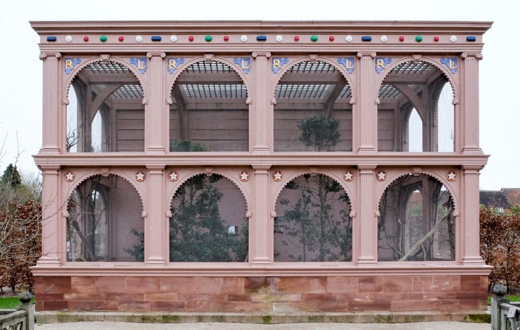 Kenilworth Castle 8 A pink-painted two-story timber aviary with round arches on both levels, the upper arches marked with gilded letters R and L, a row of red, green, blue and white painted jewels along the cornice, and trees and shrubs inside the mesh.