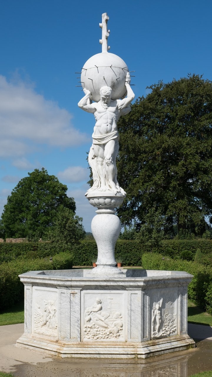 A white marble fountain with a muscular bearded figure bearing a large globe on his shoulders atop a baluster column, rising from an eight-sided basin whose panels are carved in relief with figures and sea creatures, against a blue sky and trees. A white marble fountain with a muscular bearded figure bearing a large globe on his shoulders atop a baluster column, rising from an eight-sided basin whose panels are carved in relief with figures and sea creatures, against a blue sky and trees.