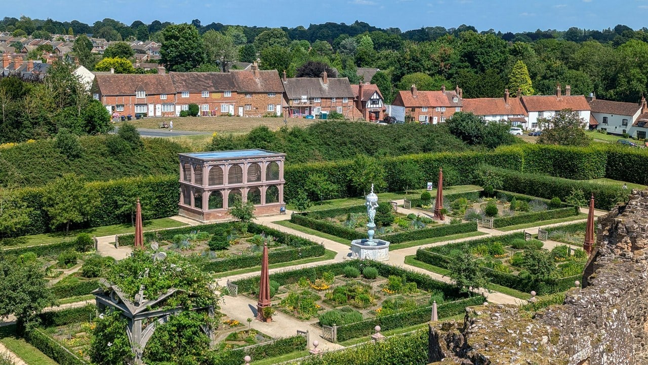 An elevated view over the Elizabethan garden: square beds edged with low lattice fencing and hedges, tall reddish obelisks, a white fountain at the center, a two-story arcaded aviary at the left, and a row of red-brick houses behind a hedge beyond. An elevated view over the Elizabethan garden: square beds edged with low lattice fencing and hedges, tall reddish obelisks, a white fountain at the center, a two-story arcaded aviary at the left, and a row of red-brick houses behind a hedge beyond.
