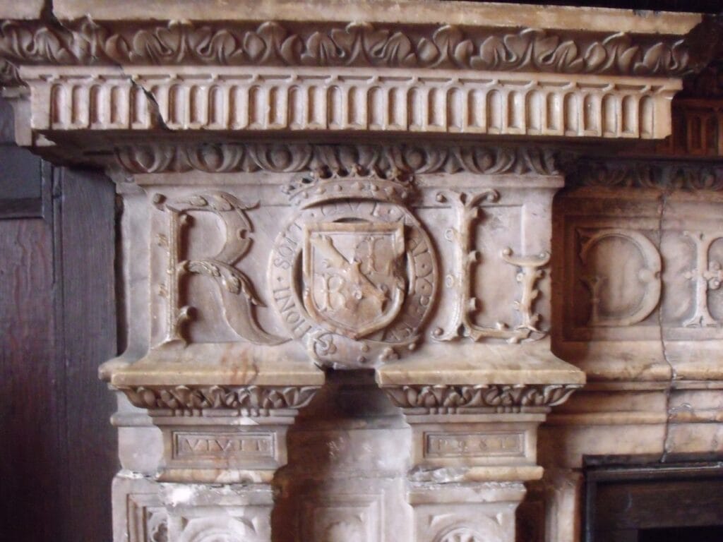 Kenilworth Castle 7 Close view of a carved pale stone fireplace: an ornate R and L on either side of a small crowned shield encircled by a garter strap, below a molded cornice with egg-and-dart and dentil ornament.