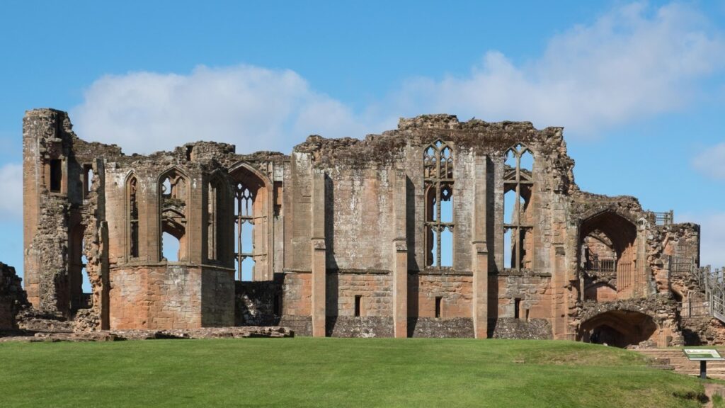 Kenilworth Castle 4 The roofless great hall range in red and gray sandstone, with tall pointed window openings with tracery, a polygonal bay at the left and a stair with railings at the right, above a mown lawn.