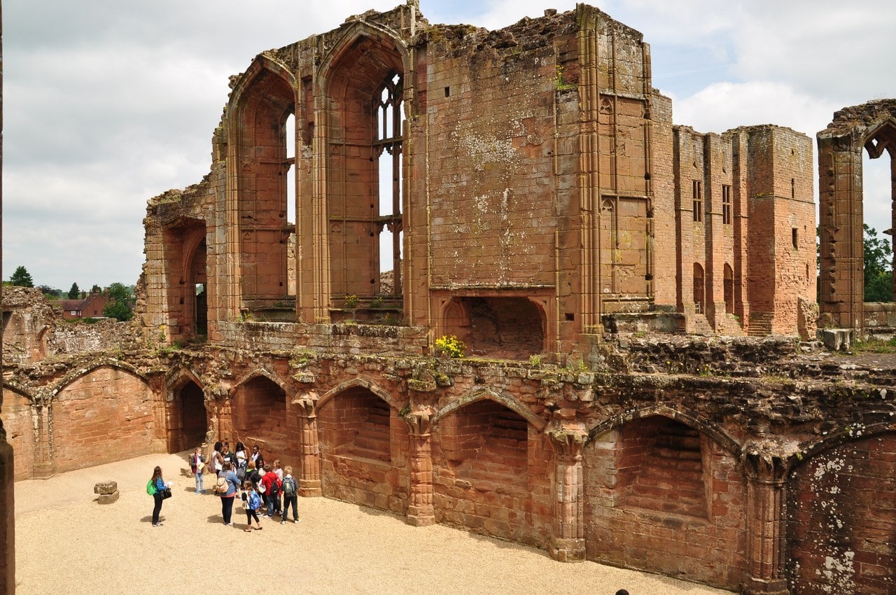The roofless red sandstone great hall seen from above, its tall pointed window openings with stone tracery and a wide fireplace opening high in the wall, and below them a row of arched undercroft bays around a gravel floor where a school group stands. The roofless red sandstone great hall seen from above, its tall pointed window openings with stone tracery and a wide fireplace opening high in the wall, and below them a row of arched undercroft bays around a gravel floor where a school group stands.