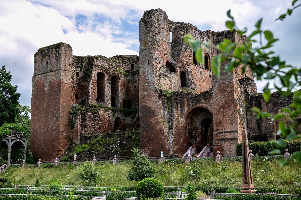 The tall red sandstone Great Tower with one side broken open to show arched openings inside, rising above a grassy bank and the planted beds of the Elizabethan garden, with a latticed arbor at the left and a slender obelisk at the right. The tall red sandstone Great Tower with one side broken open to show arched openings inside, rising above a grassy bank and the planted beds of the Elizabethan garden, with a latticed arbor at the left and a slender obelisk at the right.