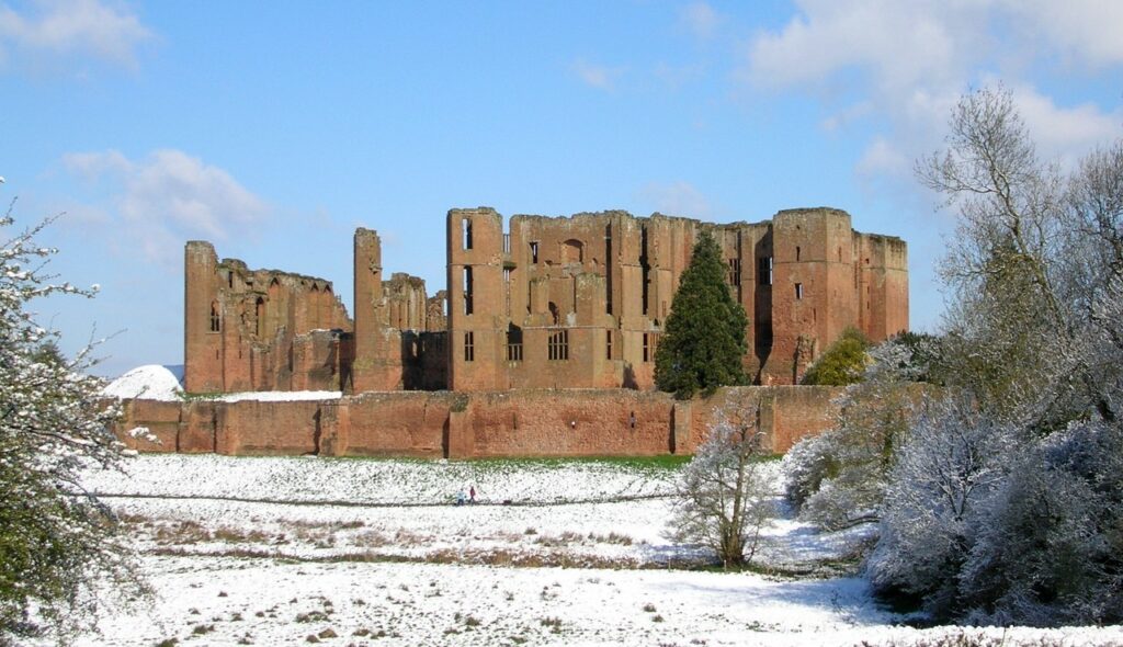 Kenilworth Castle 11 The red sandstone ruins of Kenilworth Castle rising behind the long outer curtain wall, above a meadow covered in snow, with frost-white trees on the right under a blue sky.
