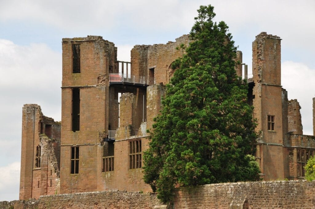 Kenilworth Castle 6 A tall ruined red sandstone building with large window openings, a modern steel walkway with railings spanning a gap near the top, and a large dark conifer in front of the right half.