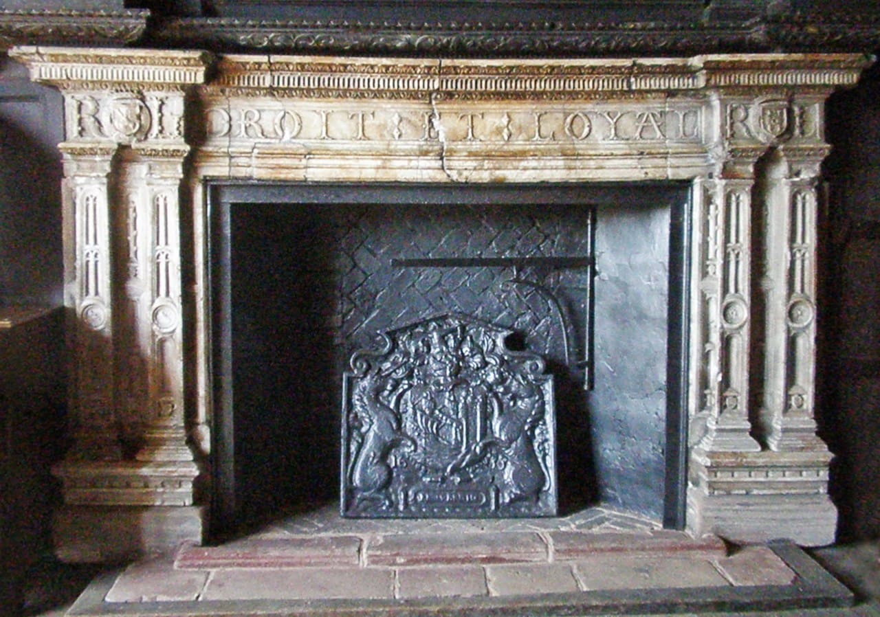 A pale carved stone fireplace with fluted classical pilasters and a frieze lettered DROIT ET LOYAL, the initials R and L carved at its end around a small shield, and a heraldic cast-iron fireback standing in the dark hearth. A pale carved stone fireplace with fluted classical pilasters and a frieze lettered DROIT ET LOYAL, the initials R and L carved at its end around a small shield, and a heraldic cast-iron fireback standing in the dark hearth.