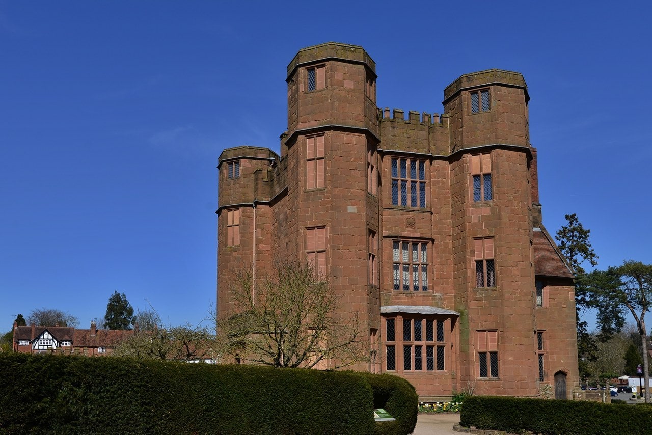 Leicester's Gatehouse, a red sandstone building with angled corner turrets, a battlemented parapet and leaded mullioned windows, seen against a clear blue sky above a clipped hedge. Leicester's Gatehouse, a red sandstone building with angled corner turrets, a battlemented parapet and leaded mullioned windows, seen against a clear blue sky above a clipped hedge.