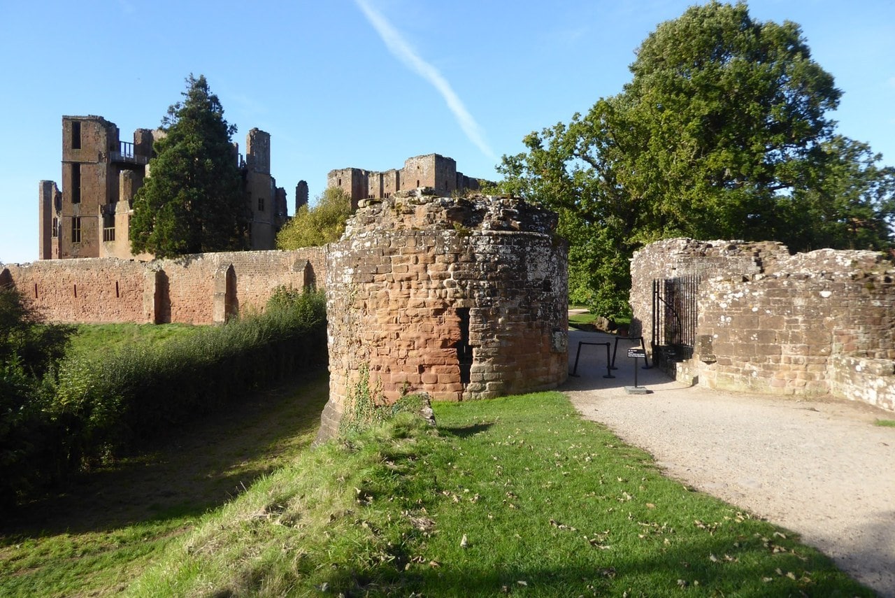 A round, ruined stump of red sandstone tower beside a gravel path and an iron gate in a low wall, with a grassy bank at the left, the castle's curtain wall behind, and tall ruins and a conifer against a blue sky. A round, ruined stump of red sandstone tower beside a gravel path and an iron gate in a low wall, with a grassy bank at the left, the castle's curtain wall behind, and tall ruins and a conifer against a blue sky.