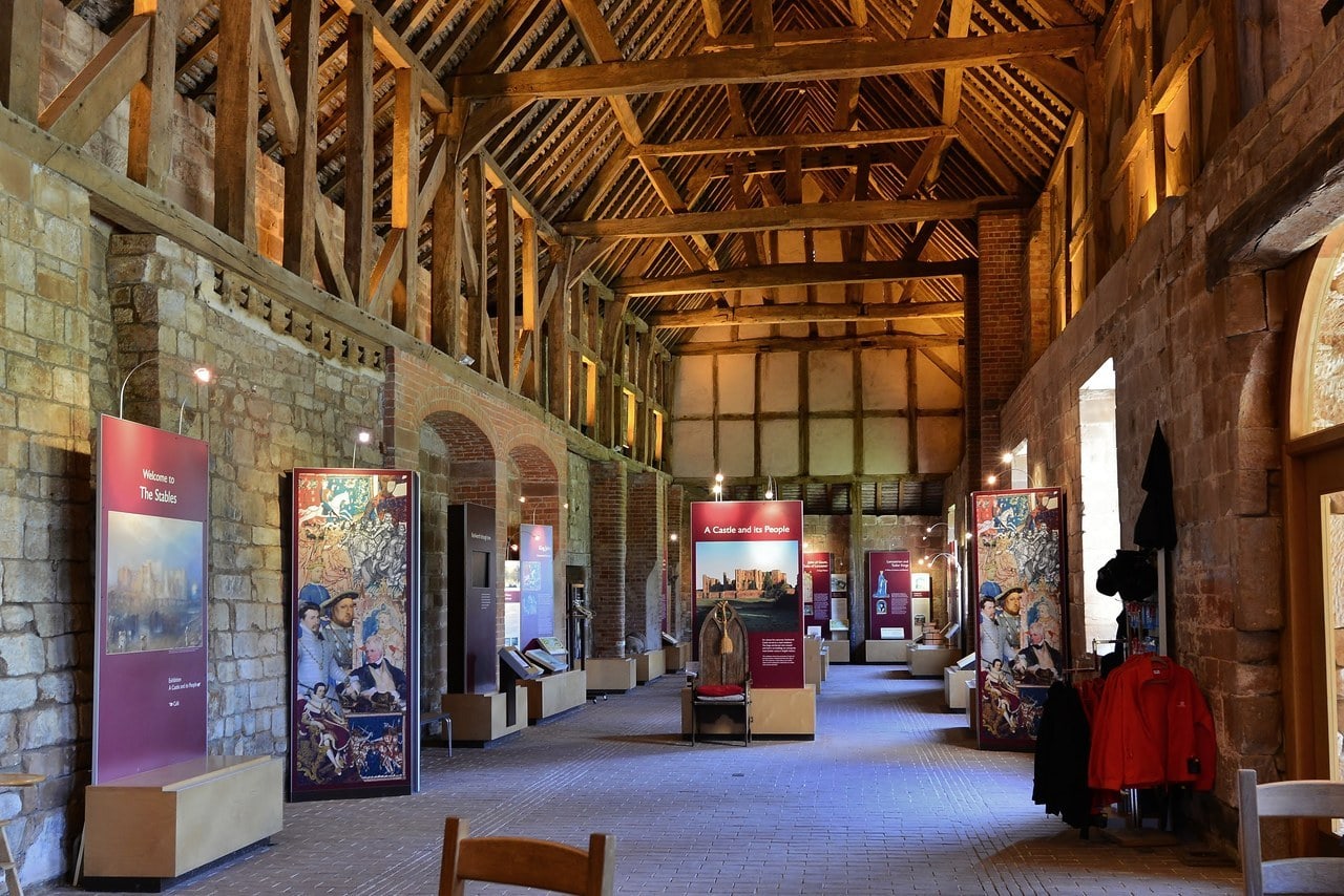 A long interior under an exposed timber roof, with stone and brick walls, a brick floor and a row of display panels and cases, the nearest panel reading Welcome to The Stables. A long interior under an exposed timber roof, with stone and brick walls, a brick floor and a row of display panels and cases, the nearest panel reading Welcome to The Stables.