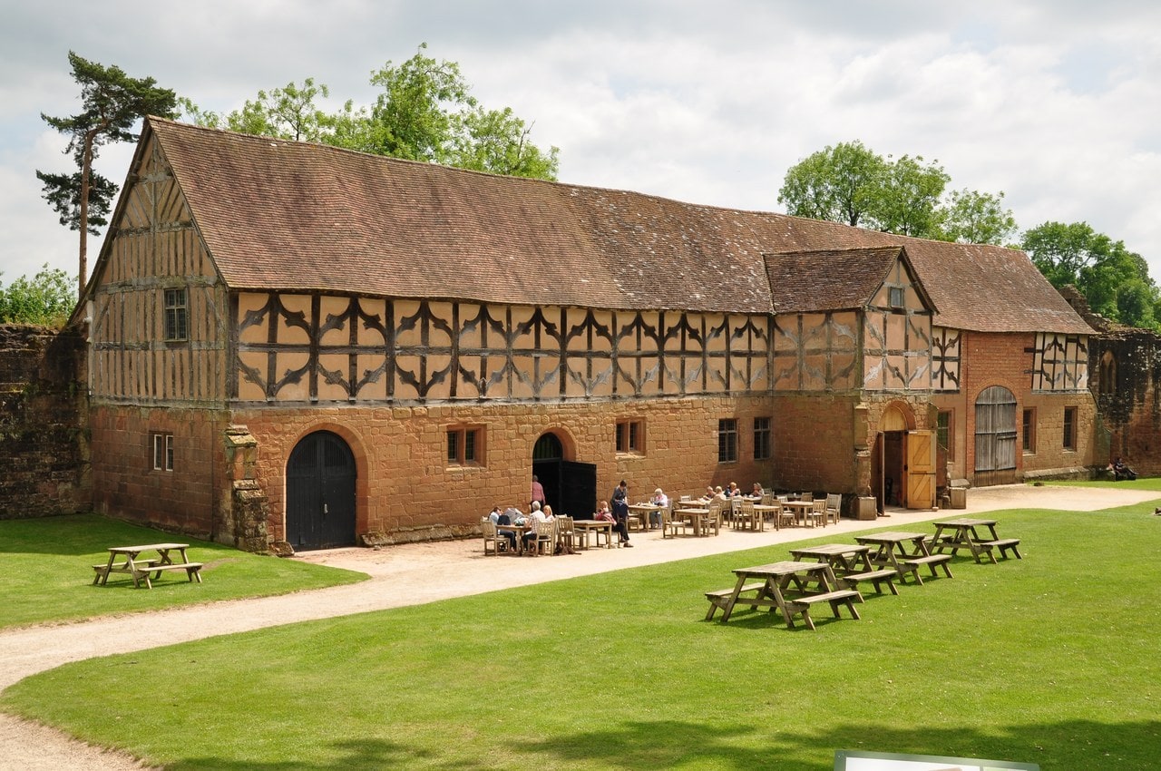 A long stable range with a red sandstone lower story and a timber-framed upper story with decorative curved braces under a steep tiled roof, with picnic tables on the lawn and visitors seated at tables along its front. A long stable range with a red sandstone lower story and a timber-framed upper story with decorative curved braces under a steep tiled roof, with picnic tables on the lawn and visitors seated at tables along its front.