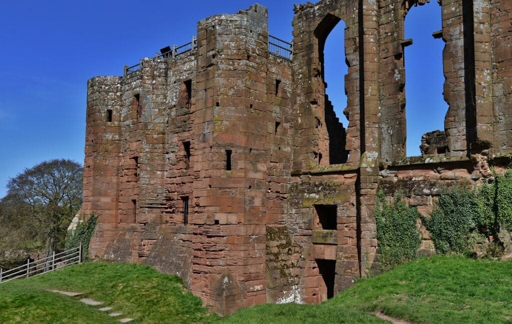 Kenilworth Castle 9 A tall, weathered red sandstone tower with small rectangular slits and a railed viewing platform on top, joined at the right to a wall pierced by very tall pointed openings, above a grassy bank.