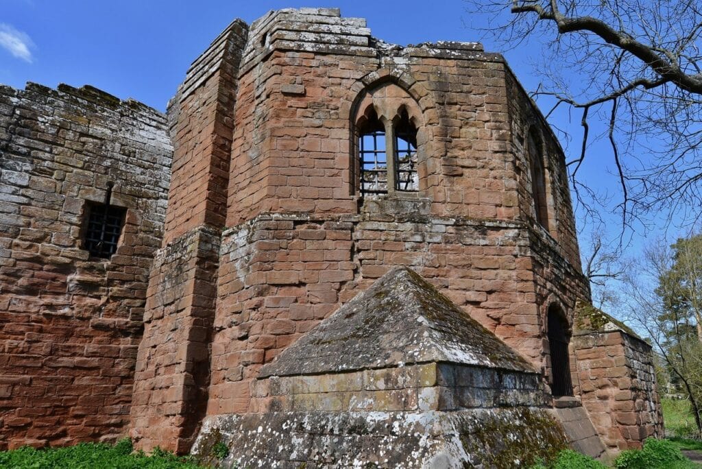 Kenilworth Castle 10 A polygonal red sandstone tower with a pointed two-light window high up and an iron-grilled doorway, with a sloping pyramidal stone spur at its base and a bare tree at the right.