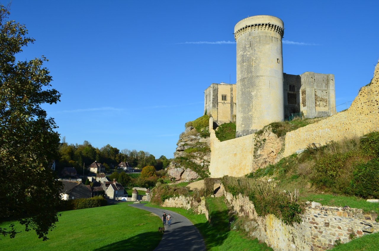 A tarmac lane curving uphill between a mown grass bank and the enclosure wall of Château de Falaise, two walkers on the lane, with the round Tour Talbot and the great tower on the rock above and village houses among trees to the left. A tarmac lane curving uphill between a mown grass bank and the enclosure wall of Château de Falaise, two walkers on the lane, with the round Tour Talbot and the great tower on the rock above and village houses among trees to the left.