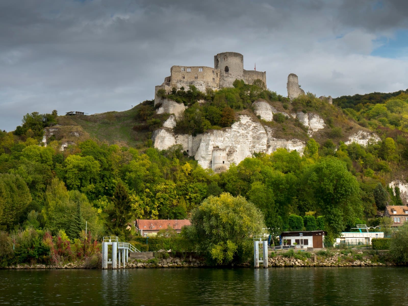 Castles in Normandy: Five Strongholds Between the Sea and the Vexin 1 Château Gaillard on a wooded chalk bluff above the Seine, the ruined keep and walls on the crest, white chalk breaking through the trees below, and a landing stage and low riverside buildings at the foot of the slope, under heavy gray cloud.