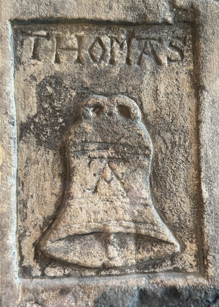 A stone panel carved with the name THOMAS above a bell bearing the letter A.
