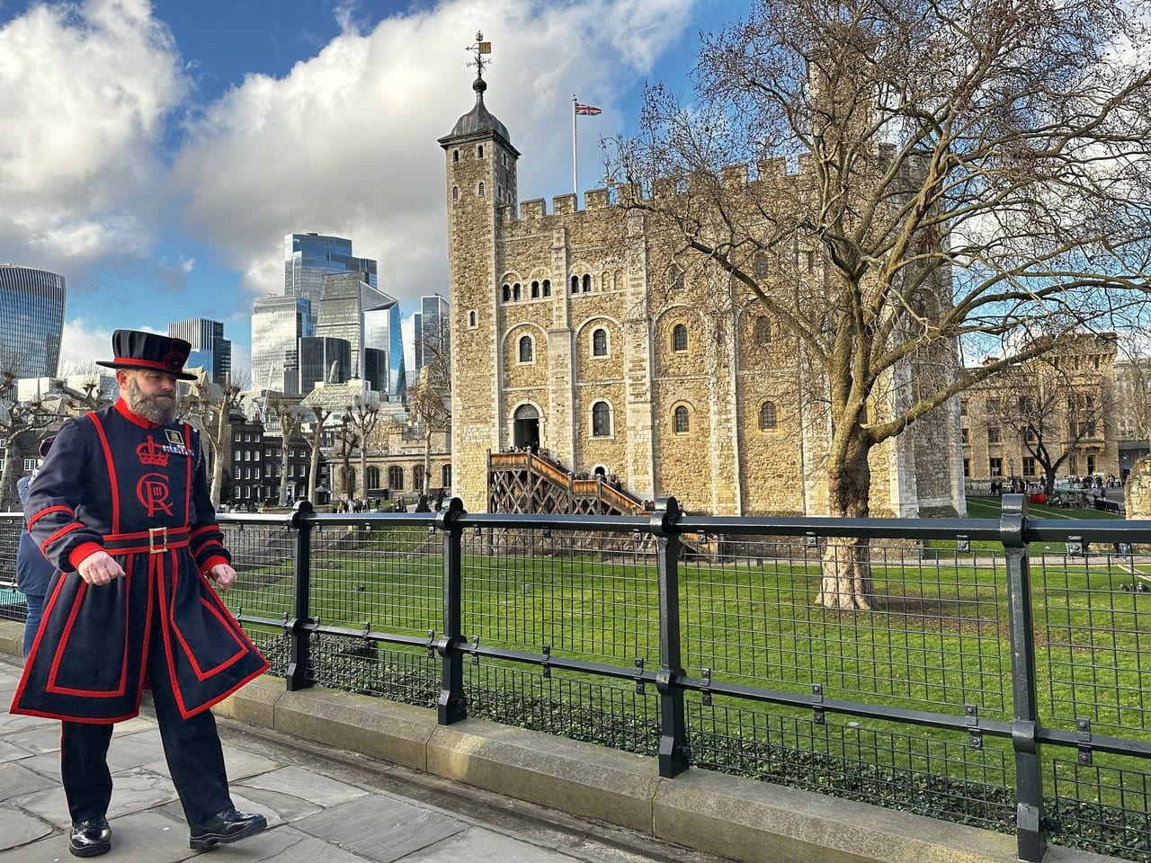 A bearded Yeoman Warder in a dark blue uniform with red trim walks along a railed path, with the White Tower and its timber stair behind.