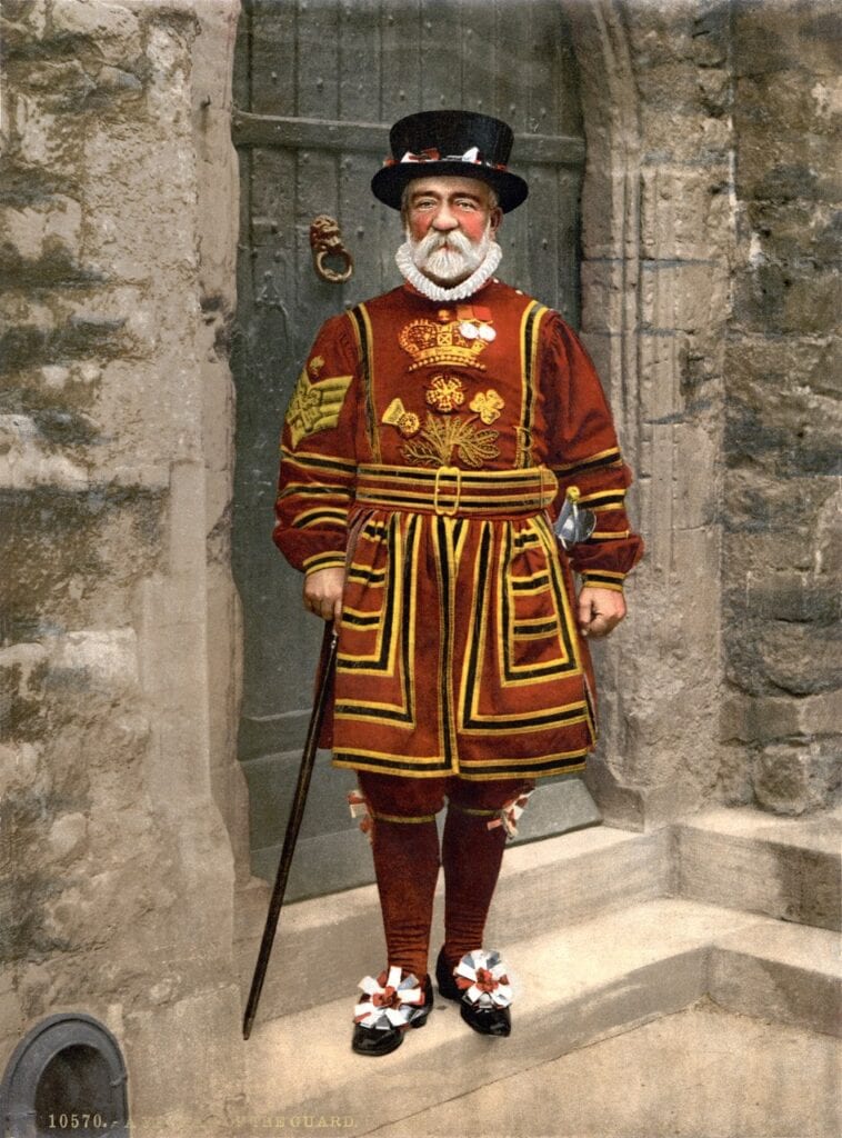 A color photograph of an elderly bearded Yeoman Warder in scarlet Tudor state dress with gold embroidery, a ruff and a black hat, holding a staff in a stone doorway.