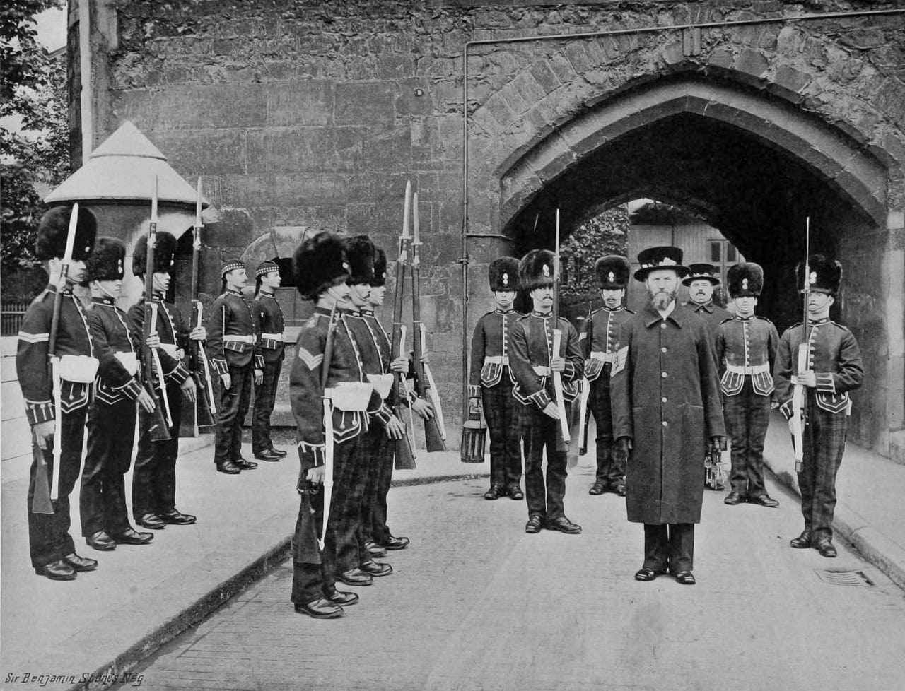 A black-and-white photograph of soldiers in fur caps with bayonets fixed, drawn up in two files before a pointed stone archway, with a bearded man in a long dark coat and Tudor bonnet standing at center right.