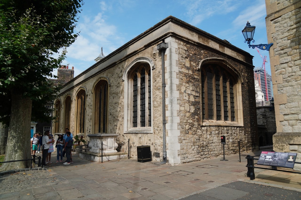 An oblique view of a rubble-built chapel with tall traceried windows and dressed stone corners, with visitors at left.