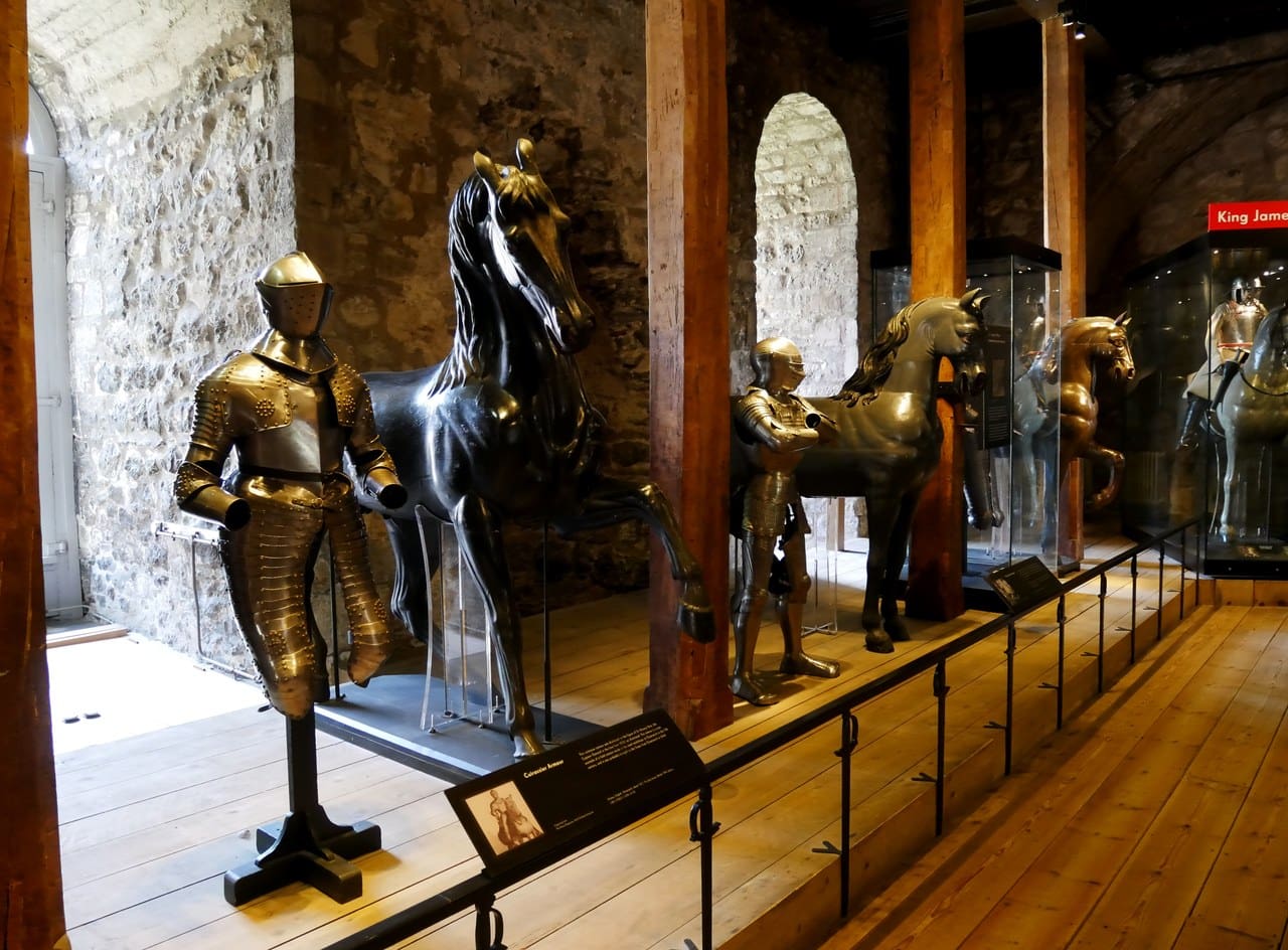 A long gallery inside the White Tower with gilded armor and carved horses in a row behind a rail, timber posts and a round-headed window.