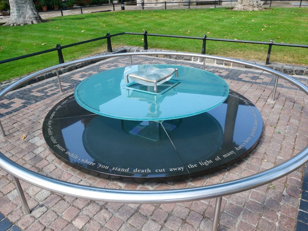 A circular glass-and-granite memorial with a glass cushion on top and an inscribed rim, behind a low steel rail, with a lawn beyond.