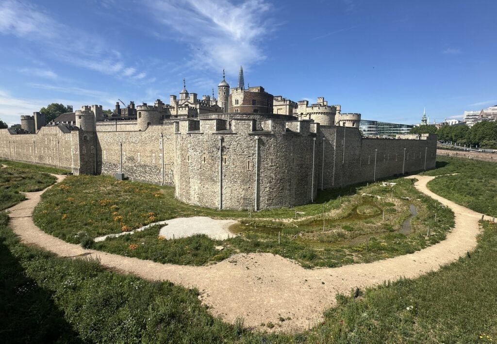 The grassed moat with a looping gravel path below the outer curtain wall as it curves to a rounded corner bastion, with towers of the inner wall behind.