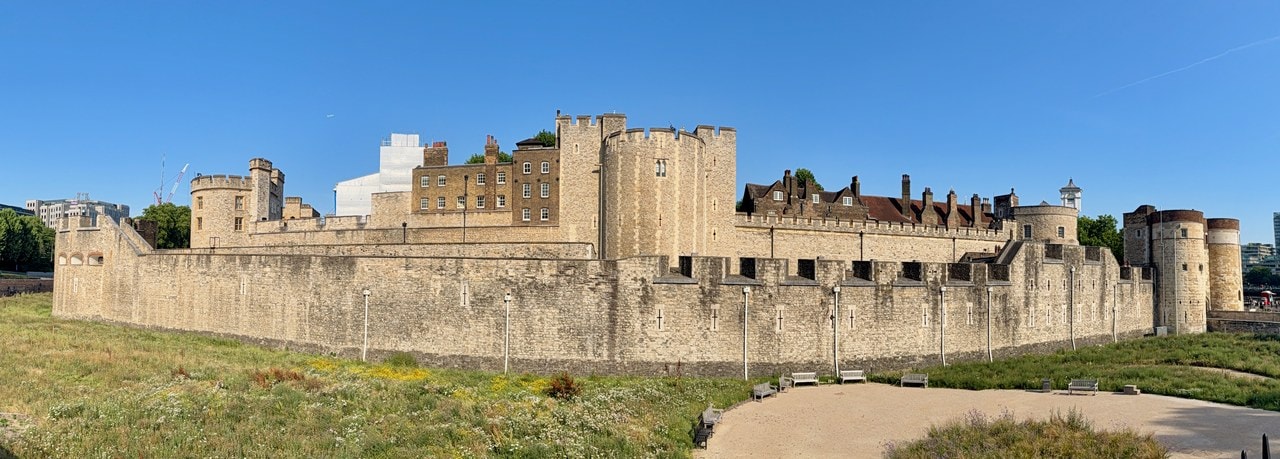 The long outer curtain wall on the Tower's west side, with towers of the inner wall behind it, seen across the dry moat, planted as a meadow with a paved path.