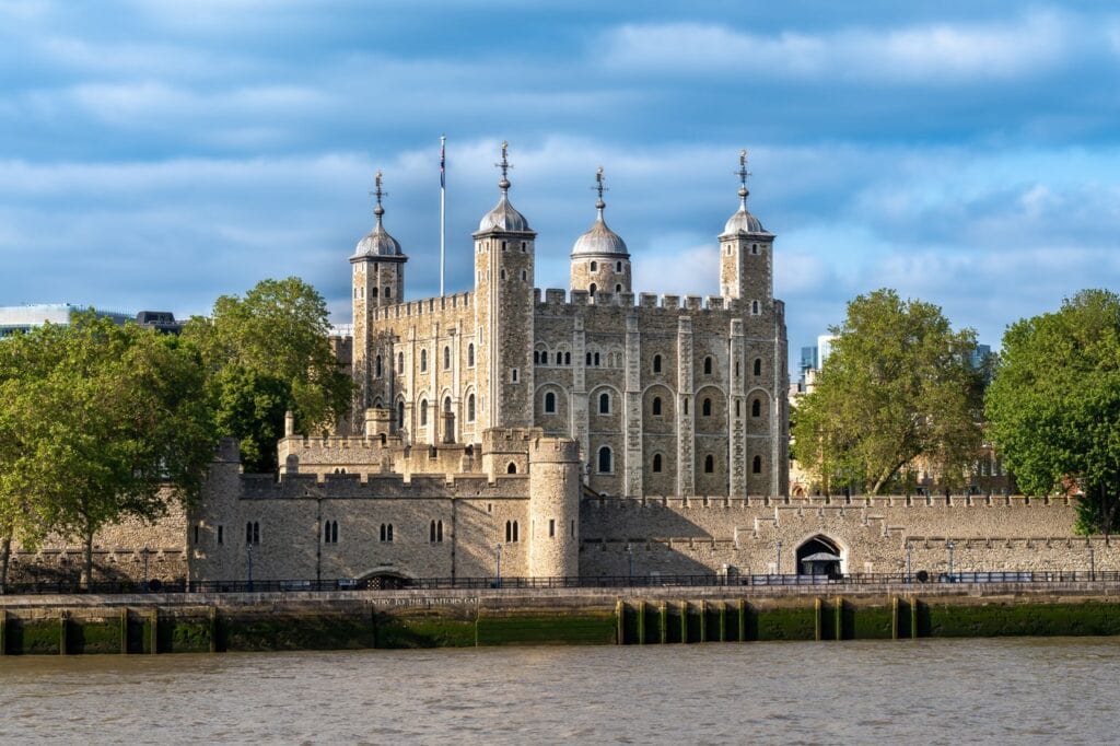 The White Tower rising above the Tower's riverside walls and trees, seen across the Thames under a cloudy blue sky.