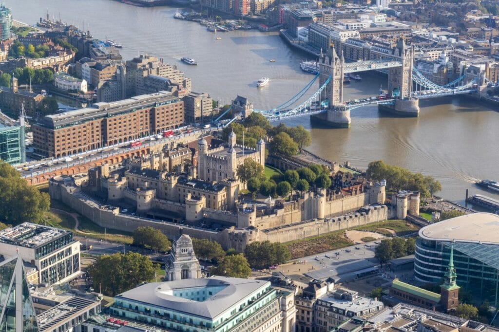 A high view over the Tower of London's walls, the White Tower and the grassed moat, with Tower Bridge and the Thames beyond.