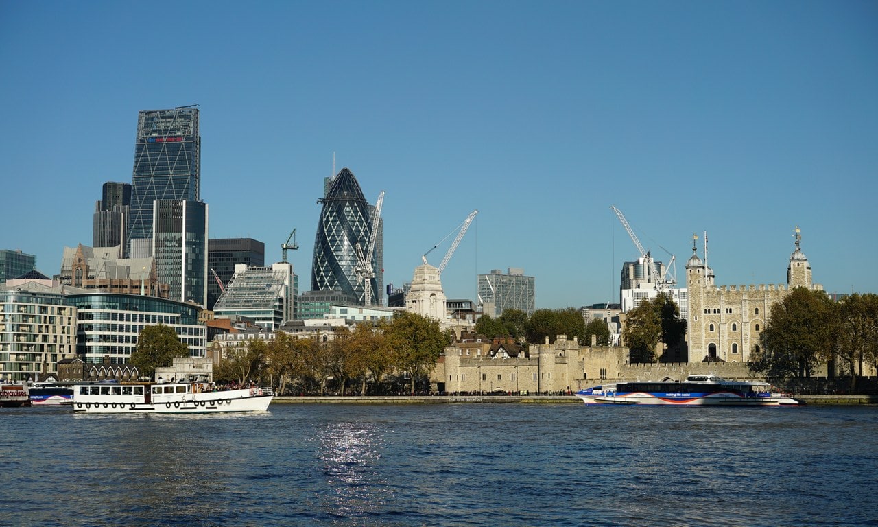 The Tower of London's riverside walls and the pale White Tower seen across the Thames, with the modern towers of the City of London rising to the left.
