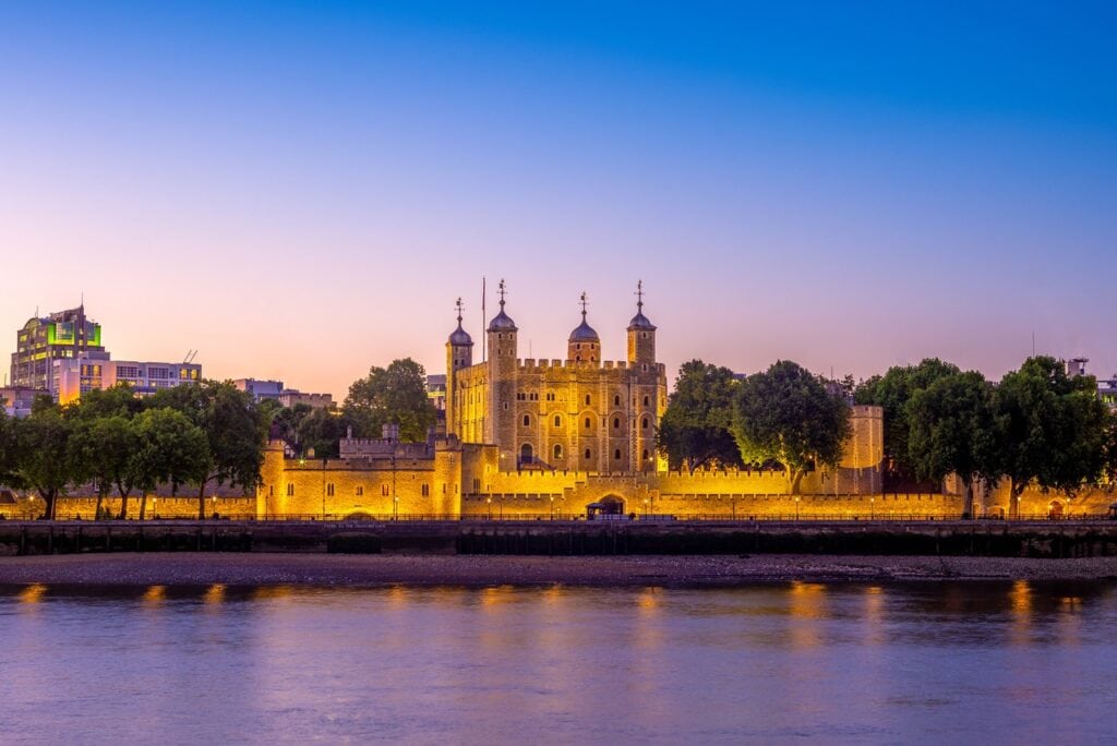 The Tower of London floodlit at dusk, seen across the Thames under a violet sky, its lights reflected in the water.
