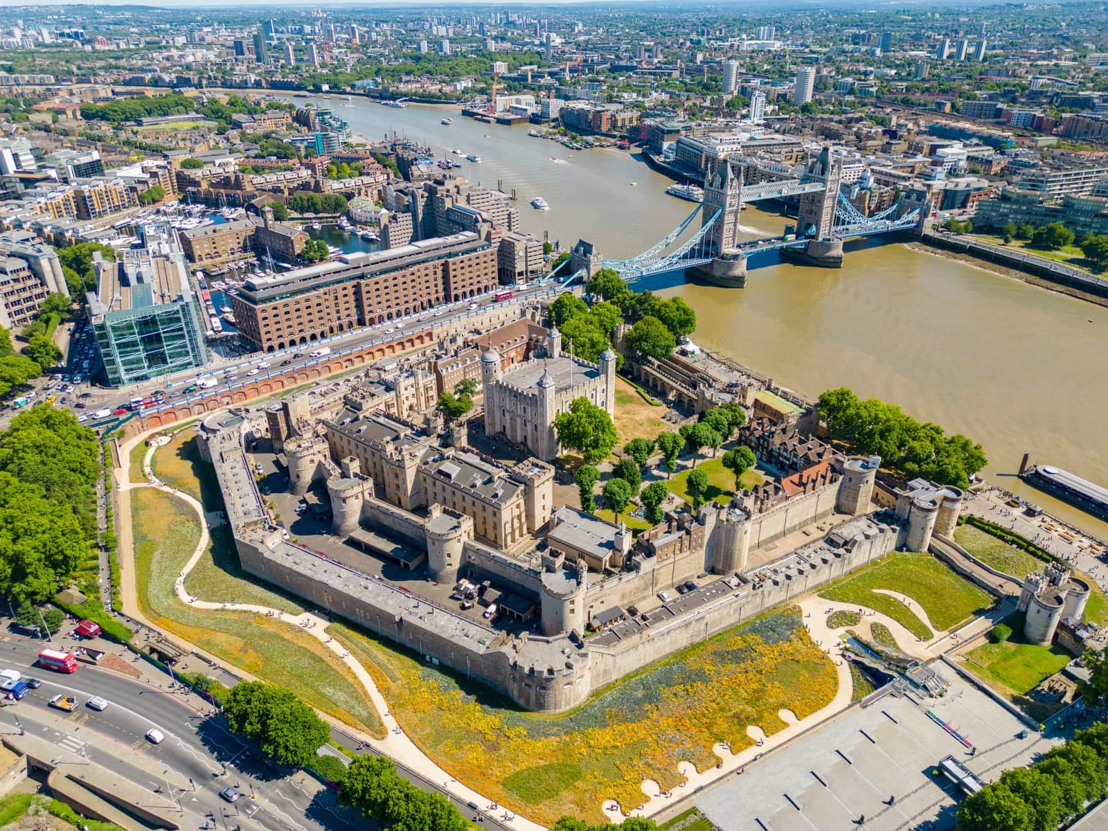 The Tower of London from the air, the White Tower at the center of its walls, wildflowers in bloom in the moat, and Tower Bridge and the Thames beyond.