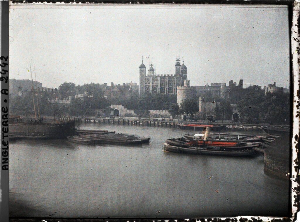 An early color photograph of the White Tower and the river frontage seen across the Thames, with a tug and barges, inside a dark plate border.
