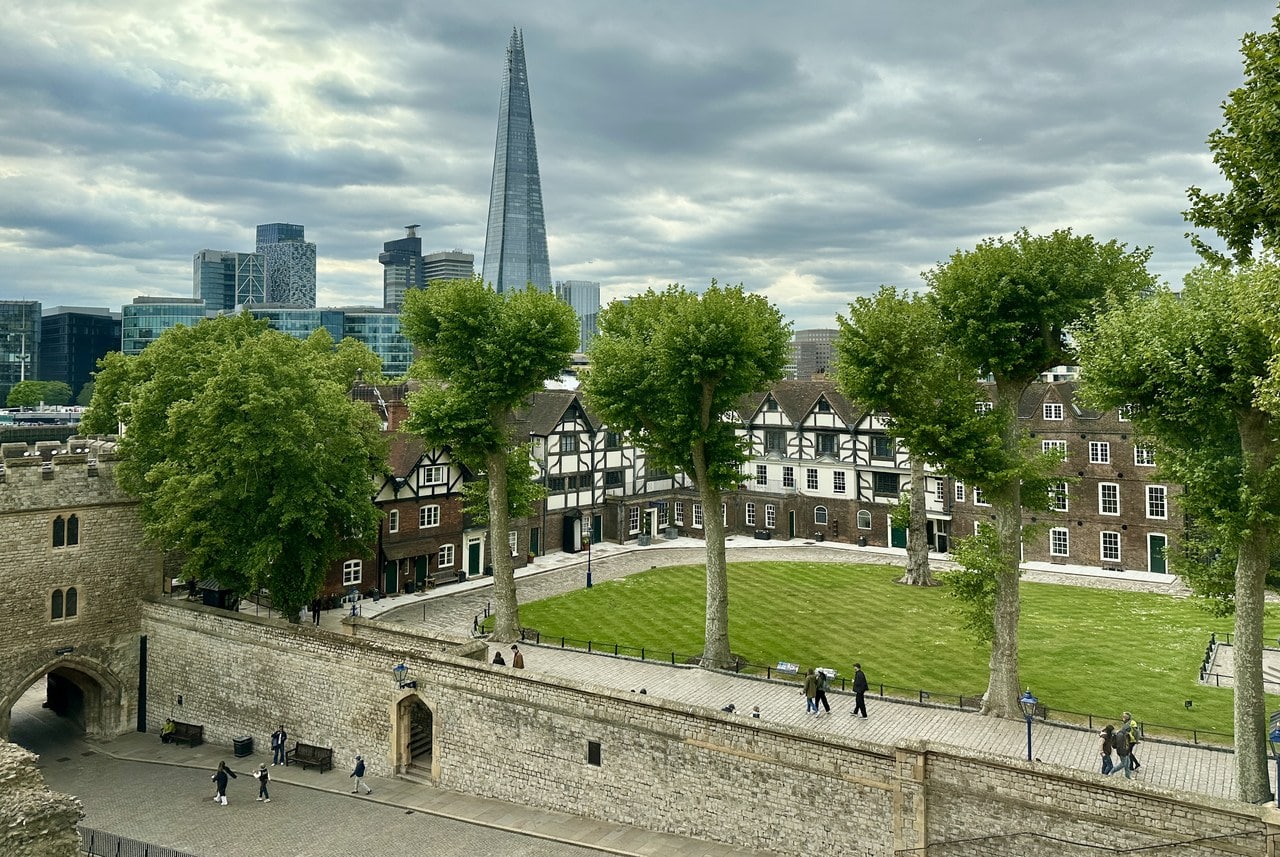 The lawn of Tower Green seen from a raised viewpoint, with the timber-framed King's House and brick houses along its far side and the Shard in the distance.