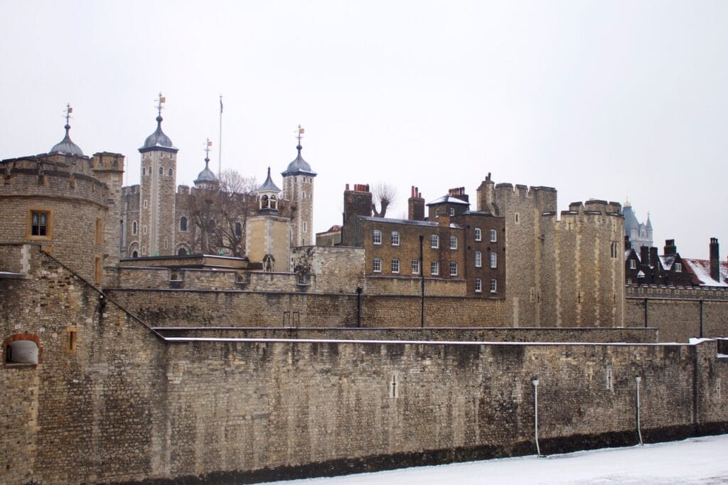 Snow on the ground below the Tower's outer wall, with the inner wall, brick houses and the White Tower's turrets behind under a white sky.