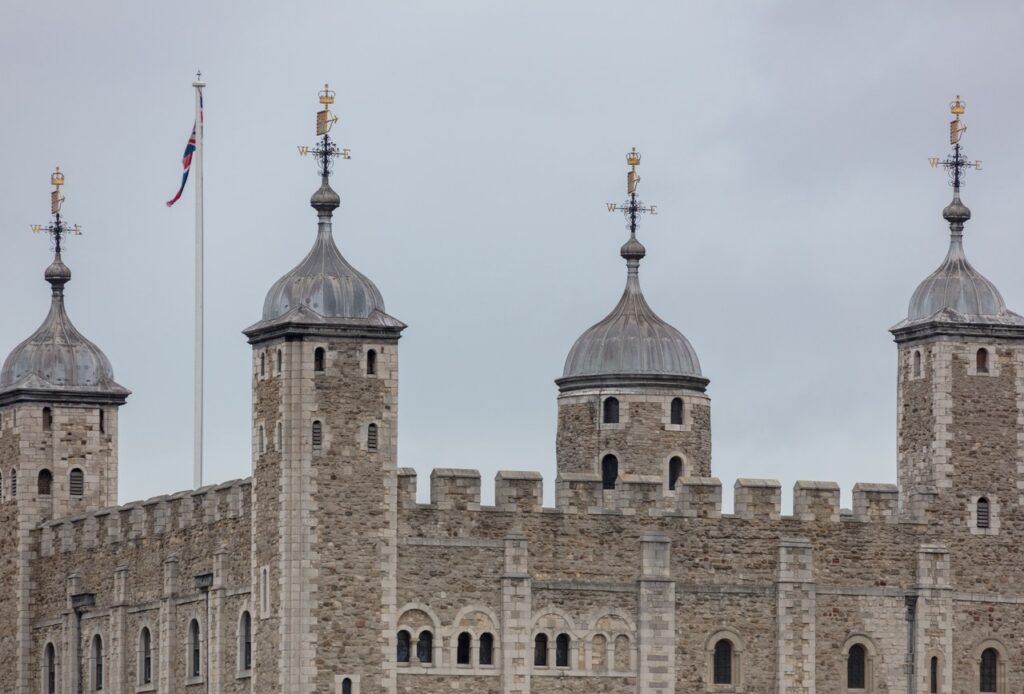 The top of the White Tower: a crenellated parapet with four turrets under domed caps and weathervanes, and a flagpole, against an overcast sky.