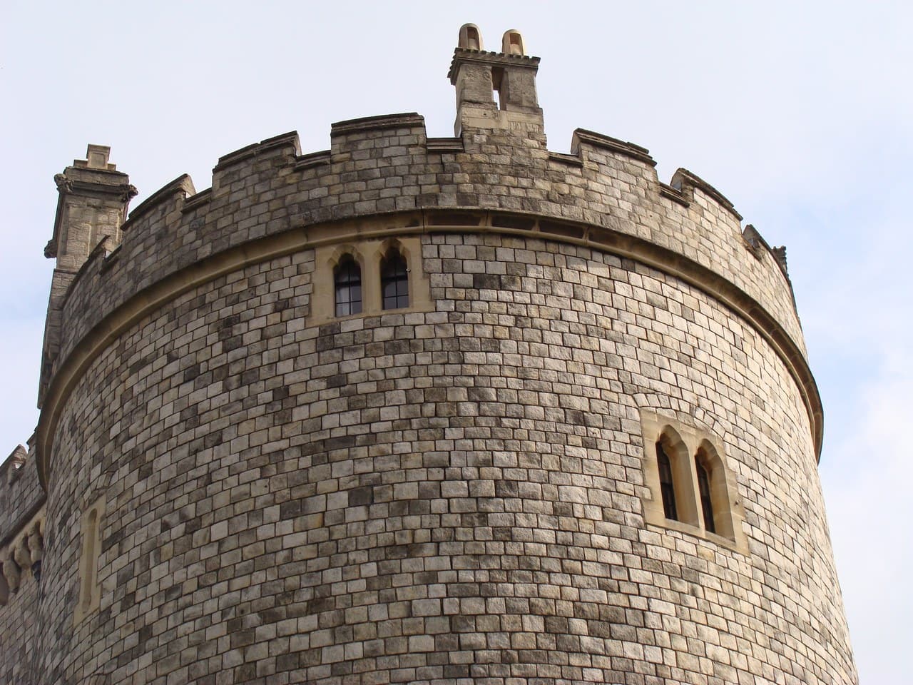 Close view of the crenellated top of a round stone tower, a molded string course below the parapet and a two-light Gothic window in the wall Close view of the crenellated top of a round stone tower, a molded string course below the parapet and a two-light Gothic window in the wall