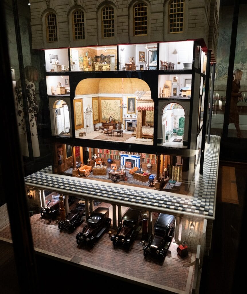 Windsor Castle 16 A large dolls' house displayed in a glass case with its front open, showing lit miniature rooms on three floors above a row of miniature motor cars