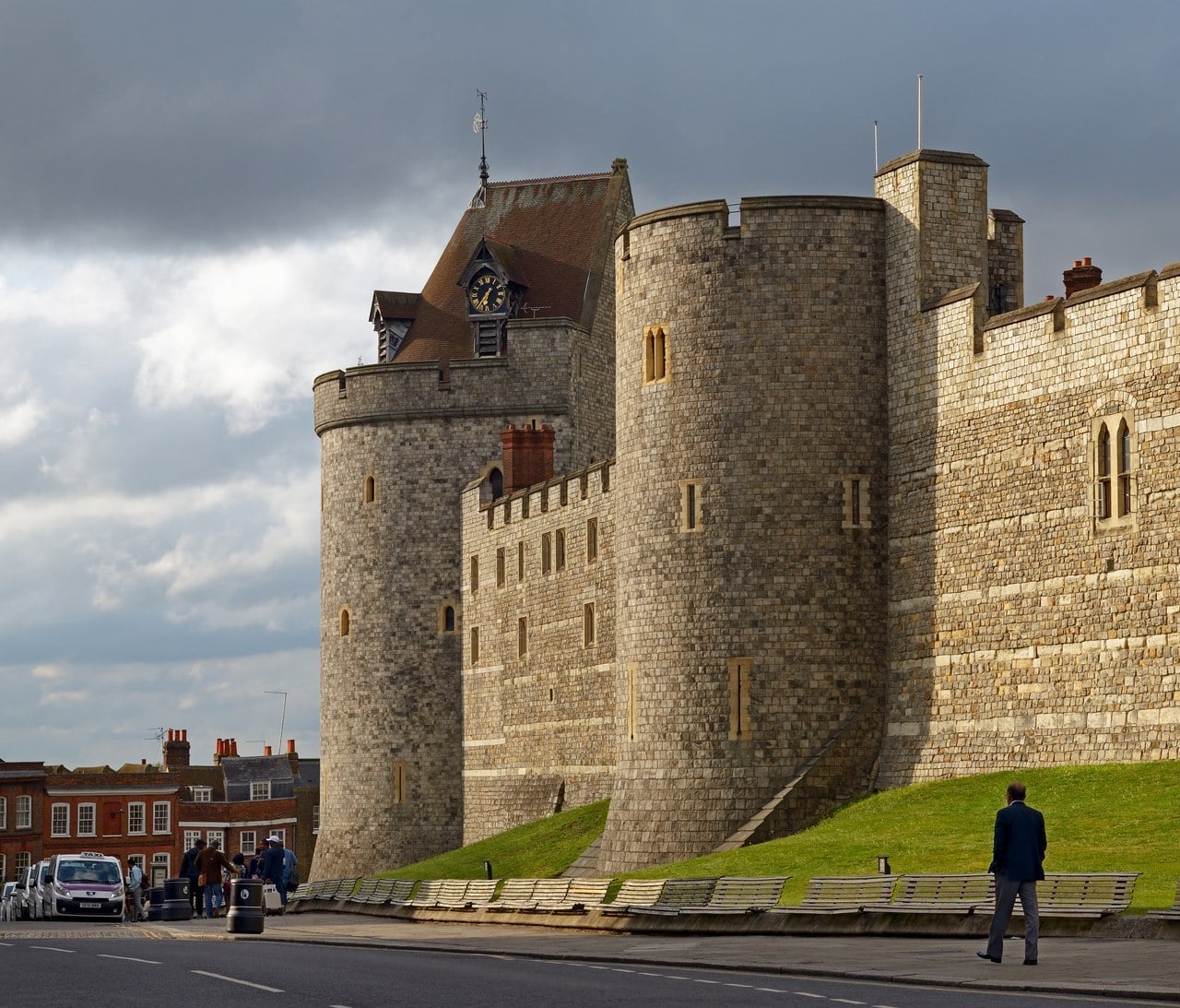 A drum tower with a steep roof and a clock at the end of a long battlemented stone wall running down a grass slope A drum tower with a steep roof and a clock at the end of a long battlemented stone wall running down a grass slope