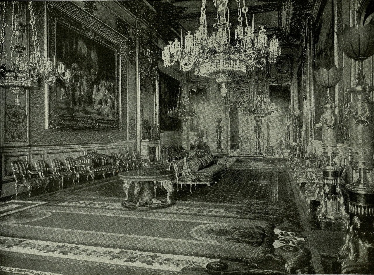 A monochrome halftone photograph of a long, heavily gilded reception room hung with tapestries, with chandeliers and gilt furniture A monochrome halftone photograph of a long, heavily gilded reception room hung with tapestries, with chandeliers and gilt furniture