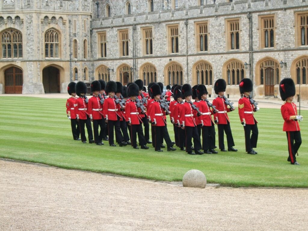 Windsor Castle 17 A rank of guardsmen in scarlet tunics and bearskin caps drawn up on a grass quadrangle, with a battlemented stone range behind them