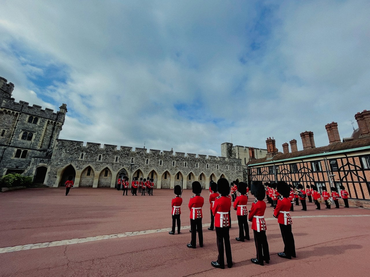 Two files of guardsmen in scarlet tunics and bearskin caps standing in a paved ward, with a battlemented stone range on one side and a timber-framed range behind Two files of guardsmen in scarlet tunics and bearskin caps standing in a paved ward, with a battlemented stone range on one side and a timber-framed range behind