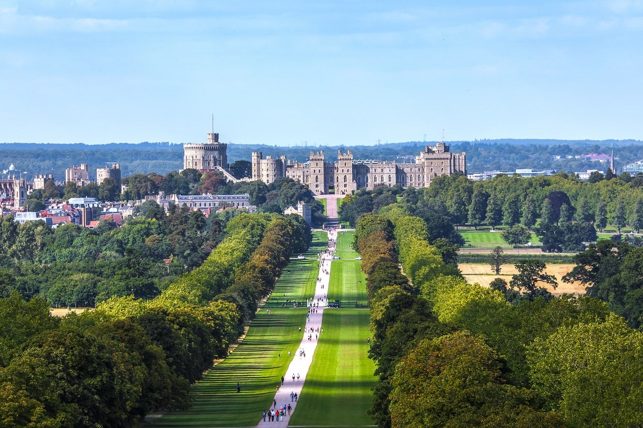 An elevated view north along a broad straight grass avenue lined with mature trees, leading to a castle on a low ridge in the far distance with a town beyond An elevated view north along a broad straight grass avenue lined with mature trees, leading to a castle on a low ridge in the far distance with a town beyond