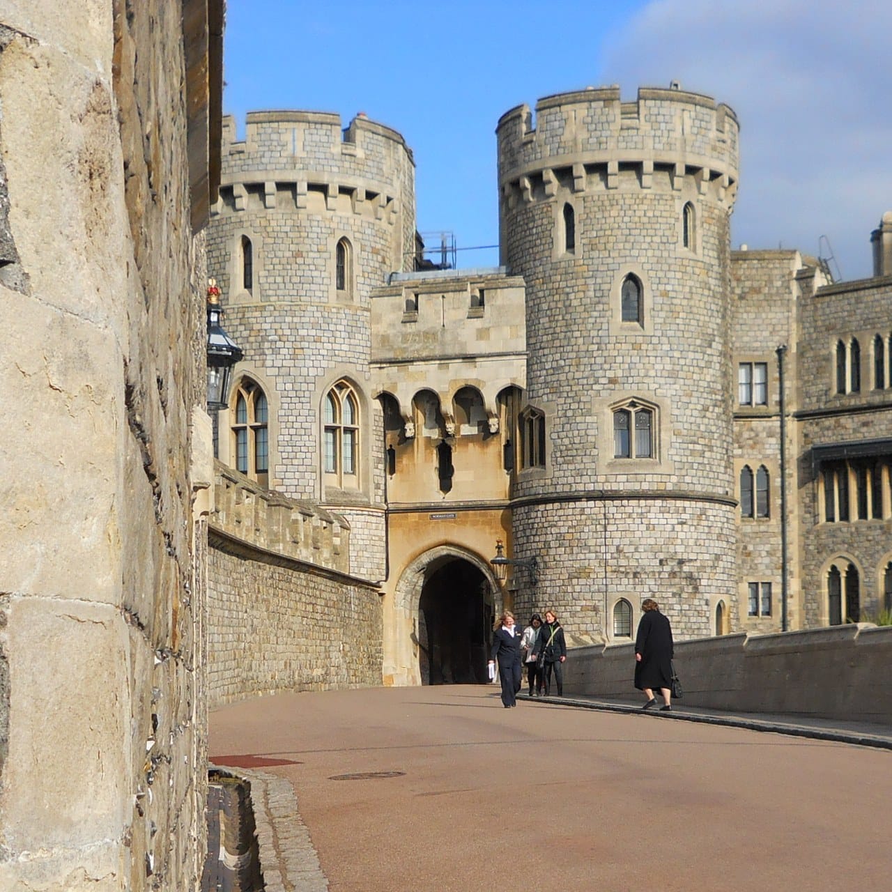 Two round stone drum towers flanking a pointed gateway, with a battlemented range behind and visitors walking up the paved approach Two round stone drum towers flanking a pointed gateway, with a battlemented range behind and visitors walking up the paved approach
