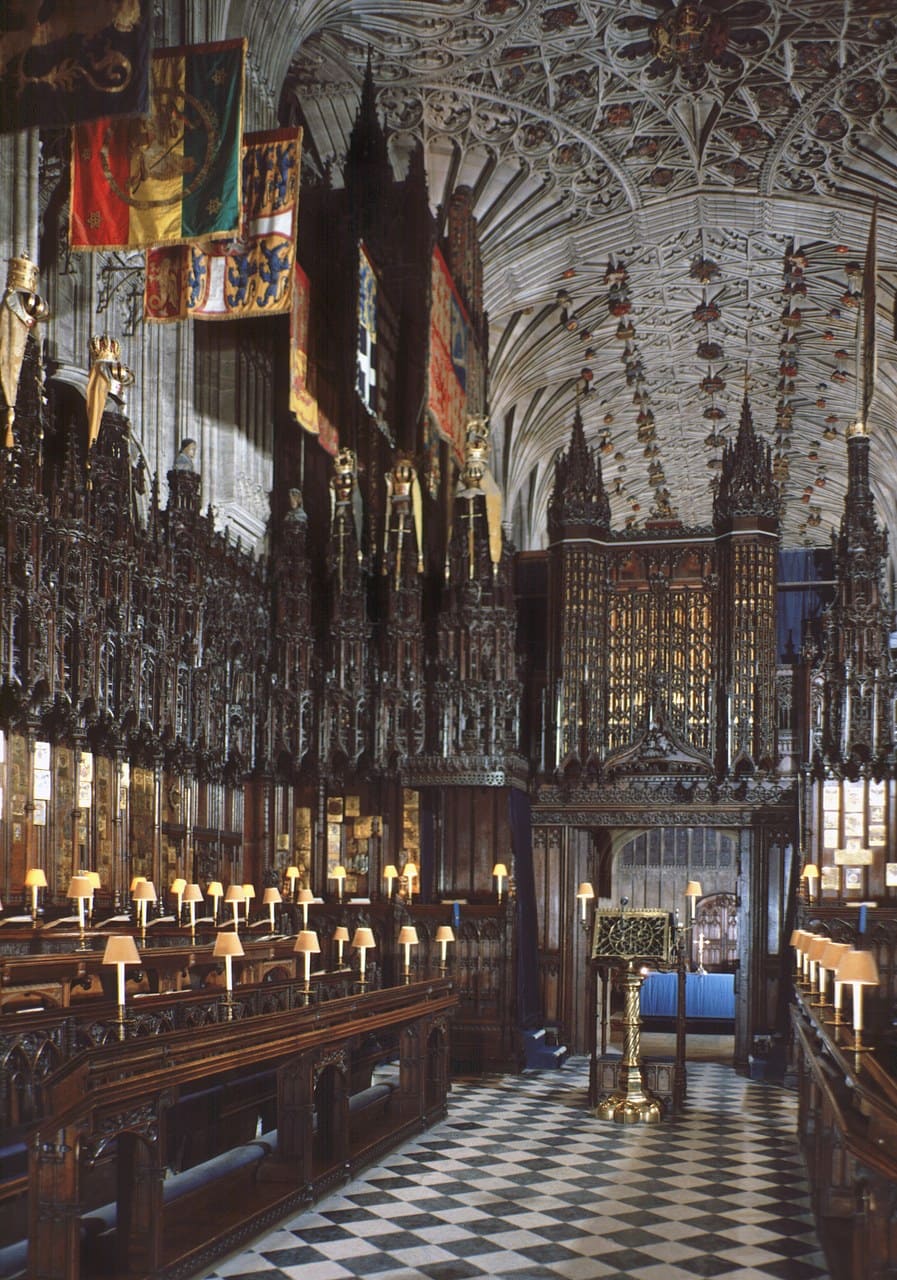 The interior of a Gothic chapel looking along two facing rows of carved wooden stalls, heraldic banners hanging above them and a vaulted ceiling overhead The interior of a Gothic chapel looking along two facing rows of carved wooden stalls, heraldic banners hanging above them and a vaulted ceiling overhead