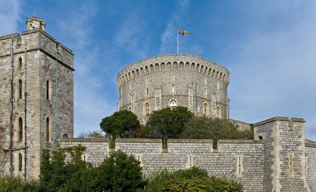 The great drum of Windsor Castle's Round Tower rising above a curtain wall and trees, a machicolated parapet round its top and a flag flying from its staff The great drum of Windsor Castle's Round Tower rising above a curtain wall and trees, a machicolated parapet round its top and a flag flying from its staff